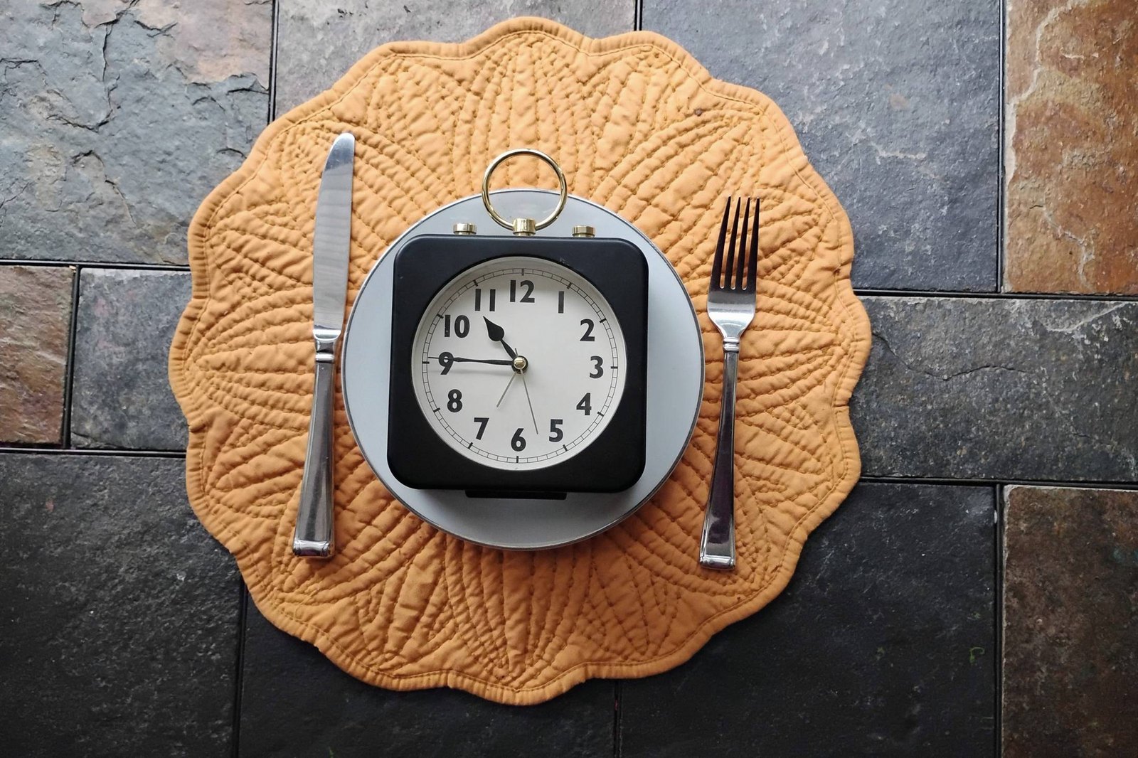 A black alarm clock on a plate between a knife and a fork, on a quilted gold placemat.