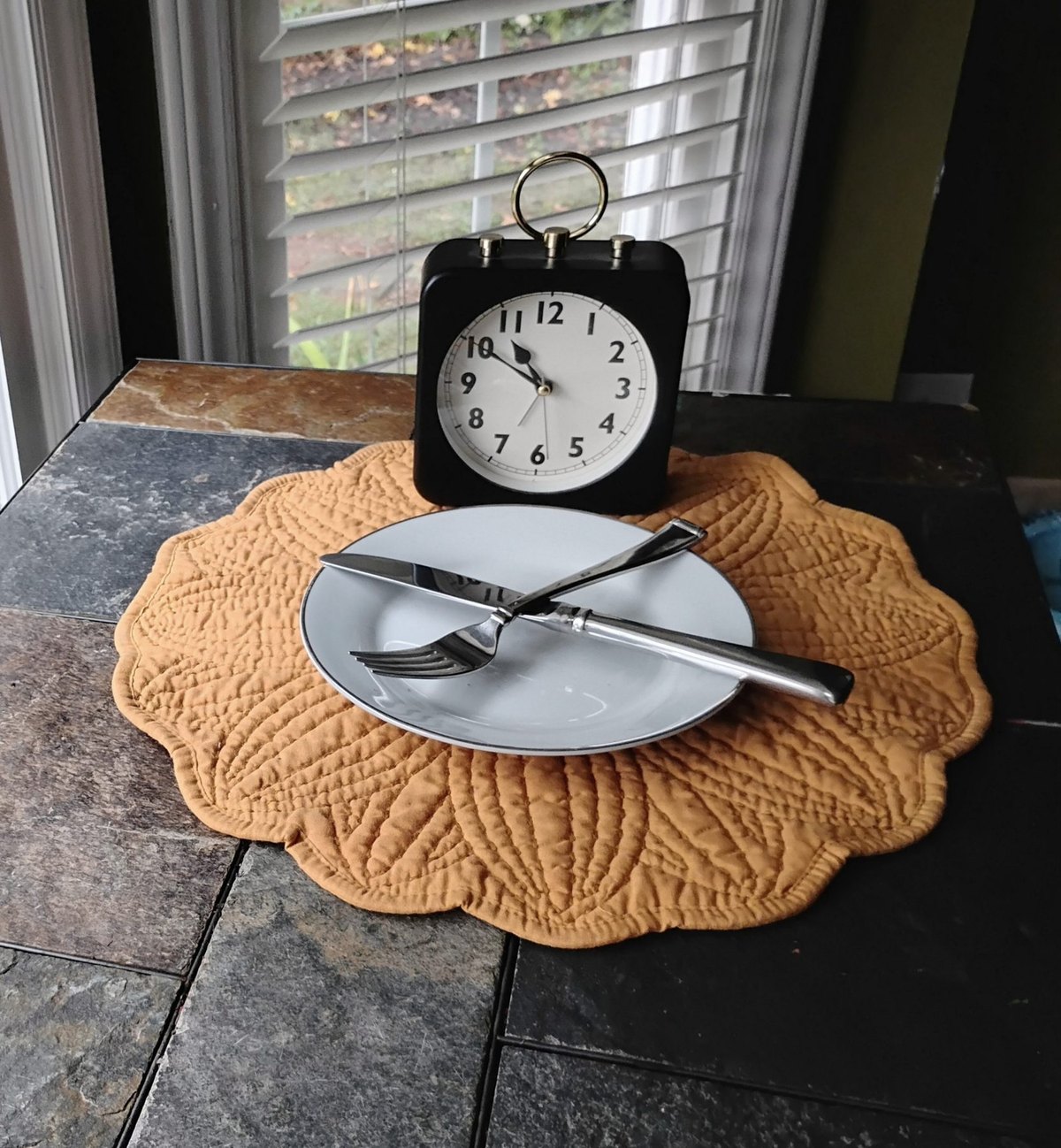 A black alarm clock behind an empty plate with a knife and fork crossed on it.