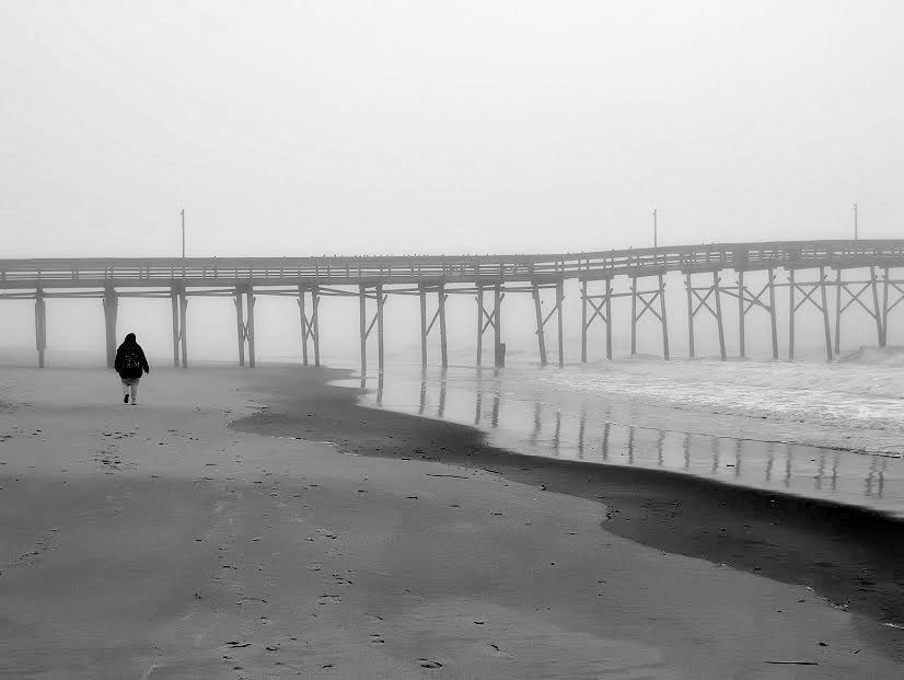 A woman walking alone on a foggy beach toward a long pier, in black and white